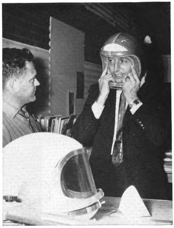 A with a helmet on his head and his hands on the helmet base as another man watches