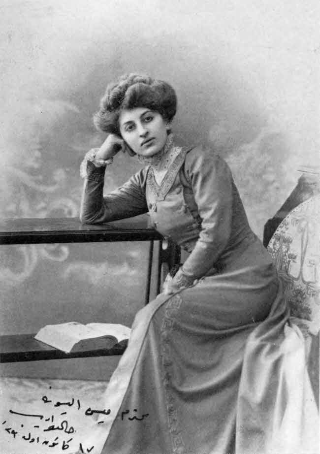 This is a formally posed almost full-body seated photo of the writer. She is leaning her left elbow on a two-tiered table with an open book on the lower shelf, with her closed hand against her cheek. She is wearing a long sleeved dress, and her hair is up but not covered. There is a hand-written inscription in Arabic script in the lower left of the photo.
