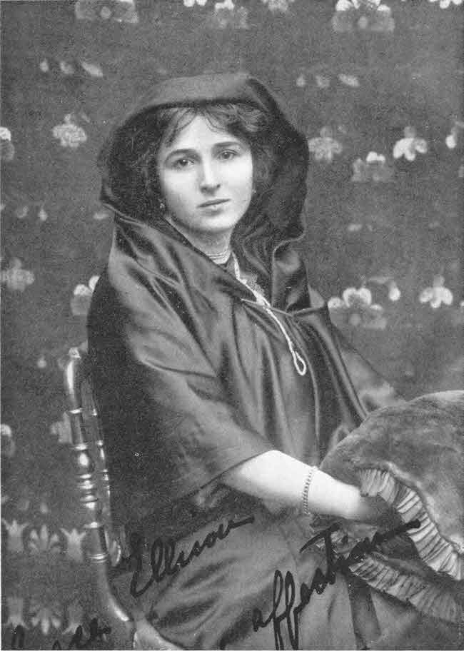 This photo shows the upper body of a seated young woman. Her body and much of her hair are covered by a flowing robe-like garment with her lower arms uncovered, and her hands under a blanket-like covering. There is a short inscription in English script.