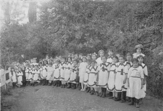 The girls are arranged standing generally in three tiers arranged in a slight semicircle receding to the left. The nearer and somewhat older-appearing girls are to the right and closer to the camera. The girls are all wearing identical white dresses with trim. Most do not have head coverings, but a few do. The girls appear to mostly be of pre-teen age. There are approximately fifty girls plus two adults, one male and one female, with one adult near each end of the semicircle.