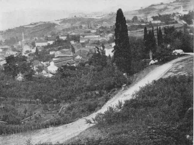 The view in this photo is from a hill overlooking the town, with what appears to be an empty dirt road nearer to the camera than the town and running through vegetation on the hillside, including trees.