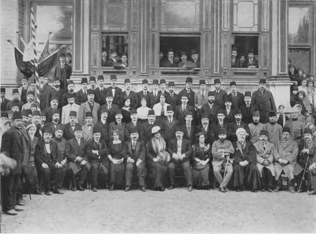 This is a formal photo of a group of adults in front of a building with windows. There are about four rows of individuals, those in front seated and those in back standing, plus some behind in open windows. A substantial majority are men, but there are some women. Almost all are formally dressed, the men generally in long coats with fez hats. There are about seventy-five people total